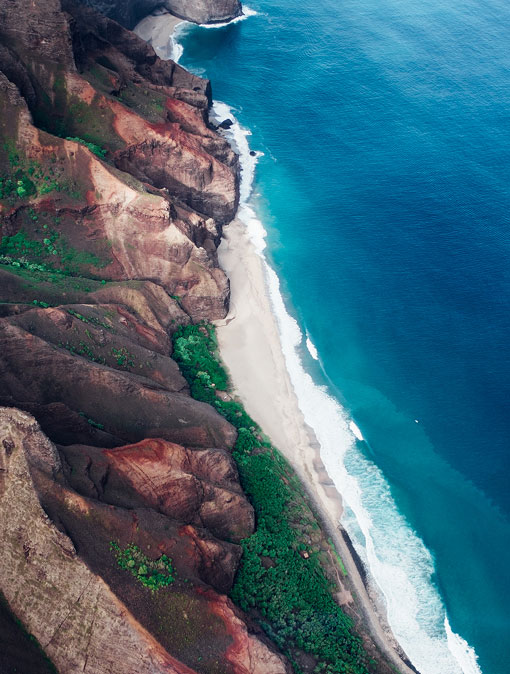 Aerial view of rugged coastal cliffs with reddish-brown rock formations, lush green patches, and turquoise waves meeting a narrow white-sand beach.